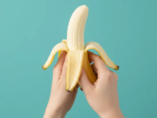 A banana being unpeeled by a pair of hands on a field of teal